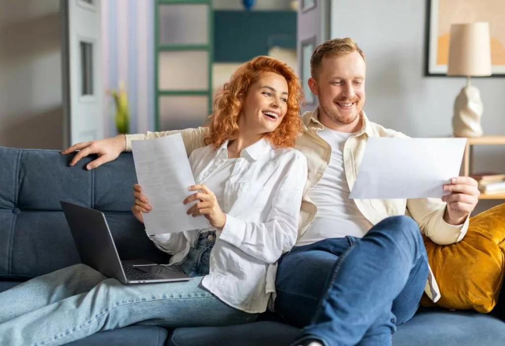 Un couple souriant consulte une attestation d’allocations familiales dans leur salon, avec des documents administratifs et un ordinateur portable à portée de main.